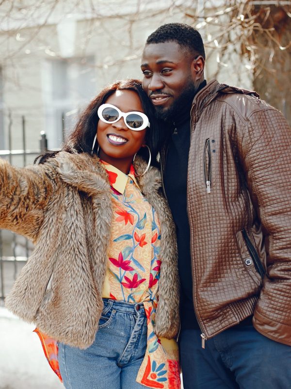 A beautiful and stylish black girl with long dark hair dressed in a brown coat and shirt standing in a summer city with her boyfriend and use the phone